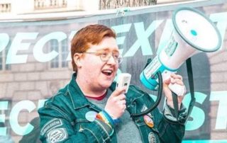Image description: young person wearing glasses holds a megaphone and wears badges representing social justice causes